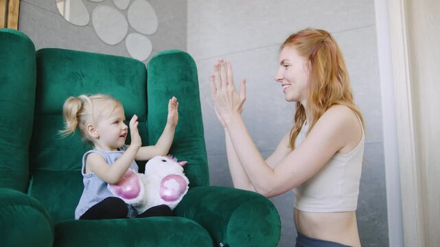 Mother and daughter share a joyful moment playing a hand-clapping game in a cozy living room with green armchair and plush toy
