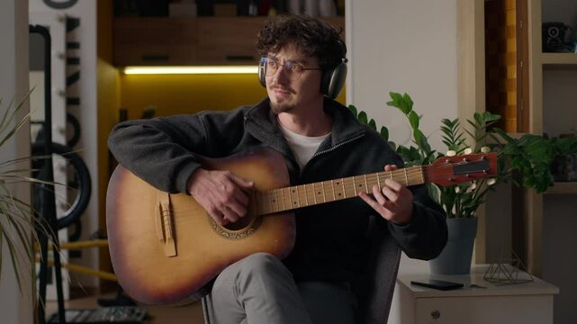 Relaxed Hispanic guitarist hipster playing acoustic guitar at home cheerful man in headphones creating melody male practicing music musician recording song guy learning play musical instrument talent