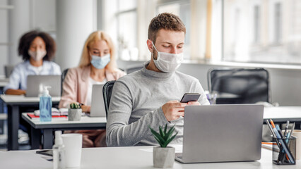 Modern technology for work during quarantine covid-19. Young man manager in protective mask typing on smartphone at workplace with laptop, plant and antiseptic in office interior with colleagues