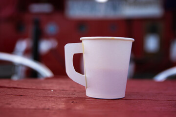 White paper coffee cup on red wooden cafe table in front of a docked red boat on blurred background.
