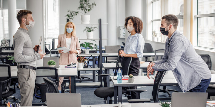 Modern team meeting, group work and social distancing. Manager with tablet speaks with workers in protective masks in interior of modern office with gadgets during coronavirus epidemic, free space - Powered by Adobe