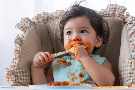 Adorable messy little child girl hungry use hand eating spaghetti sitting in high-powered chair at home. Toddler child dirty with tomato sauce making mess face looking at food. Self-feeding concept.