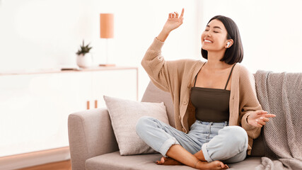 Obraz premium Music Lover Concept. Portrait of smiling asian woman wearing wireless earbuds listening to favorite song, sitting on sofa in living room. Happy millennial lady resting on couch and dancing