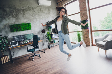 Young joyful female programmer jumping in modern home office workspace with computers and green...