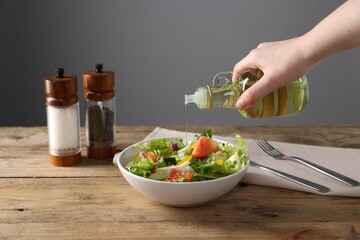 Woman pouring oil onto salad at wooden table against grey background, closeup