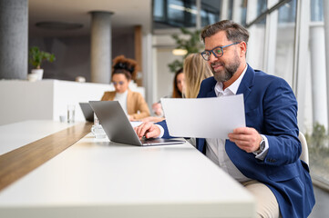 Businessman analyzing document while working on laptop in modern office with female coworkers in the background