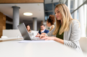 Focused blond businesswoman analyzing data over laptop while colleagues collaborating in the...
