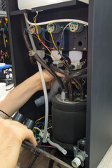 Technician performing maintenance on internal components of commercial coffee machine with wiring, hoses, and hydraulic system exposed

