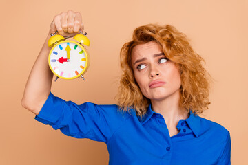 Young professional woman holding alarm clock with a playful expression in a studio. Vibrant blue shirt and red hair