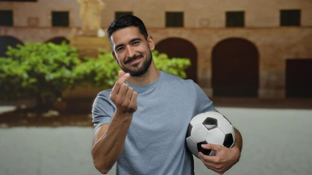 Young man making heart gesture with fingers while holding a soccer ball on a city street showing a playful and cheerful expression.
