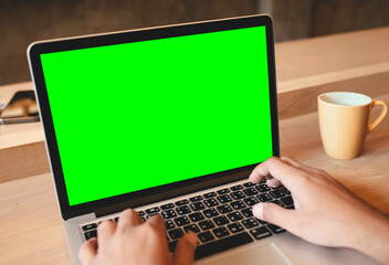 Fototapeta premium Person typing on laptop with green screen at wooden desk with coffee mug in warm indoor environment.