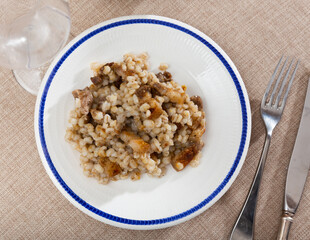 on burlap texture tablecloth there is plate with blue edge with portion of stewed barley porridge with fried bacon, complemented by glass of red wine