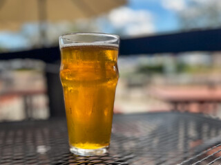 A pint glass filled with a pale ale craft beer. The liquid has a yellow tint. A clear beer glass sits on a table at a microbrewery. The background is patio umbrellas and patio tables on a sunny day.