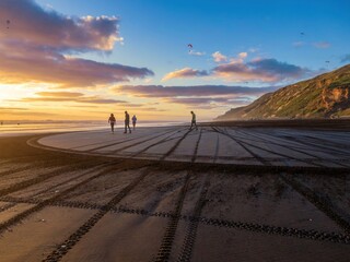 People walk and enjoy paragliding at sunset on Karioitahi Beach, Waiuku, Auckland, New Zealand. The black sand beach shows tire tracks from vehicles driving on the beach.