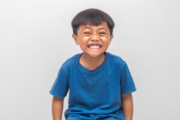 A young boy with dark hair and a blue shirt grins intensely, showcasing his teeth against a plain...