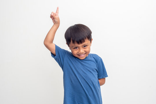 A young boy with dark hair and a blue shirt smiles while pointing his index finger upwards against a clean, white backdrop. Image toned and noise added, soft focus