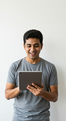 A young man with a gray shirt smiling while holding a tablet in front of a white background wall