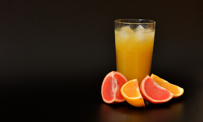 Orange and grapefruit juice in a tall cut glass with ice on a black background, next to pieces of ripe fruit.