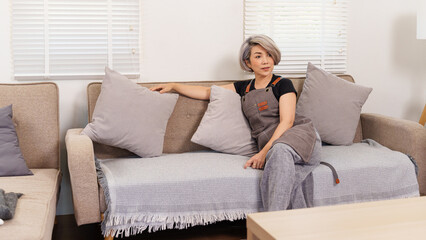 Fototapeta premium Asian Woman Relaxing on Sofa in Living Room with Apron Looking Away Interior Design Home Decor Comfort Casual Lifestyle Couch Pillows Window Blinds Natural Light