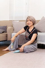 Obraz premium Older Asian Woman Smiling Holding Brush Dustpan Sitting on Floor in Living Room House Cleaning Concept Housewife Cleaning House Professional Cleaner Service at Home