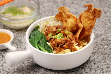a bowl of Mie Ayam, a noodle dish, topped with chicken, vegetables, and fried wontons, with a side of broth