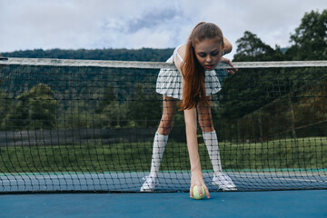 Young female tennis player in a skirt leaning down to pick up a tennis ball on the court, green...