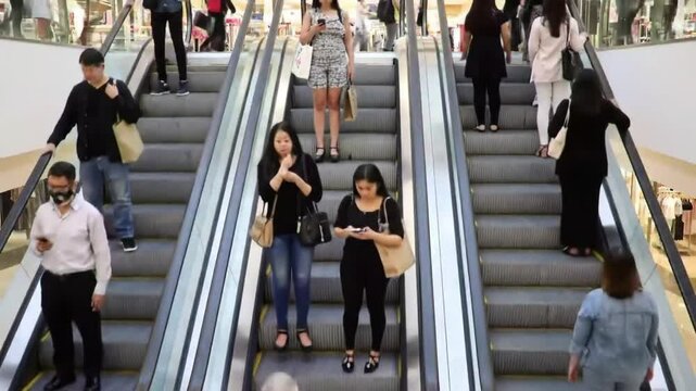 Crowds of shoppers are seen using escalators in a vibrant mall. Some visitors read, while others engage with their mobile devices, showcasing the lively atmosphere of the shopping district.