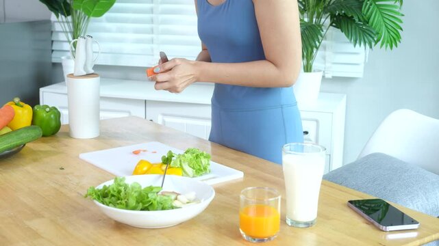 Beautiful Asian woman in fitness sportswear happily smiles while slicing vegetables and fruits on cutting board.She focuses on health nutrition at home,offering online guidance through her computer