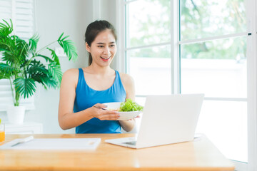 Beautiful Asian woman in fitness sportswear happily smiles while slicing vegetables and fruits on cutting board.She focuses on health nutrition at home,offering online guidance through her computer