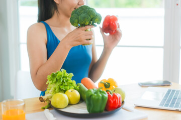 Beautiful Asian woman in fitness sportswear happily smiles while slicing vegetables and fruits on cutting board.She focuses on health nutrition at home,offering online guidance through her computer
