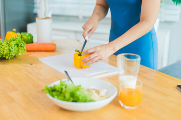 Beautiful Asian woman in fitness sportswear happily smiles while slicing vegetables and fruits on cutting board.She focuses on health nutrition at home,offering online guidance through her computer