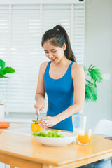 Beautiful Asian woman in fitness sportswear happily smiles while slicing vegetables and fruits on cutting board.She focuses on health nutrition at home,offering online guidance through her computer