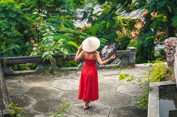 back of beautiful woman tourist traveler in a Vietnamese non la hat with a fan in a garden in...