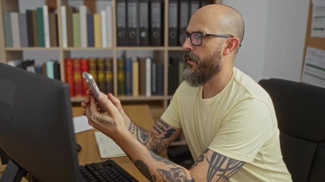Hispanic man with tattoos working in an office and checking his smartphone while sitting at a desk with a computer and surrounded by bookshelves