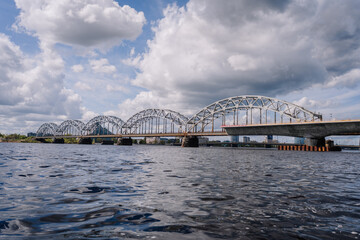 Arched railway bridge spanning across a wide river under dramatic clouds, captured from water level with reflections on the surface..