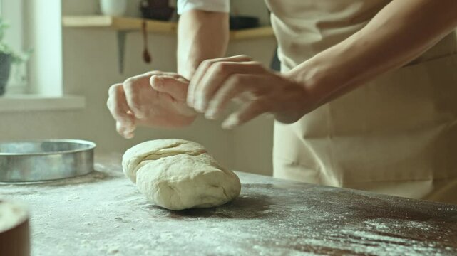 A baker in an apron kneads the dough with his hands and sprinkles it with flour to bake delicious homemade bread.