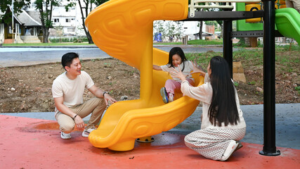 A family is standing in front of a slide. The child is sitting on the slide while the parents look...