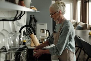 Senior woman with gray hair washed dishes in contemporary kitchen with minimalistic design. Warm light filled the room, providing serene atmosphere