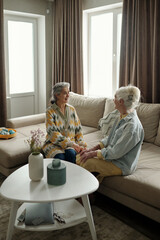 Elderly women sitting on a comfortable sofa, holding hands, in a cozy and inviting living room with natural light coming through the windows, having an intimate conversation