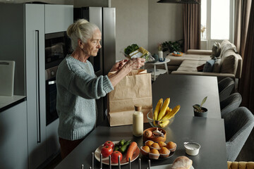 Elderly woman with grey hair unpacking groceries in a modern kitchen with various food items on the counter including fruits, vegetables, and bottled items