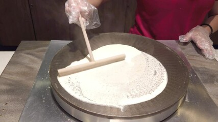 Gloved hands evenly spread crepe batter in a circular motion on a round griddle using a wooden spreader