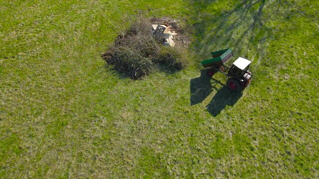 Aerial drone footage of a small vintage and retro beautiful looking tractor working in a green grassy farm field in the countryside. The tractor vehicle is carrying branches and leaves, sunny day time