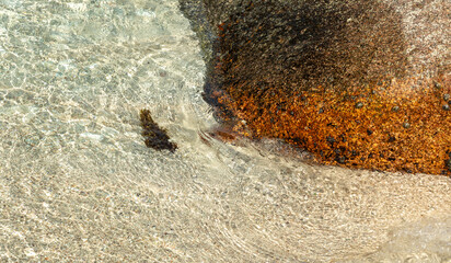 Clear ocean water flows over textured sand and orange granite rock