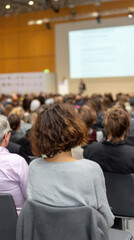 Obraz premium A woman with curly hair listens intently to a webinar speaker at a conference, with a diverse audience of attendees engaged and focused on the presentation