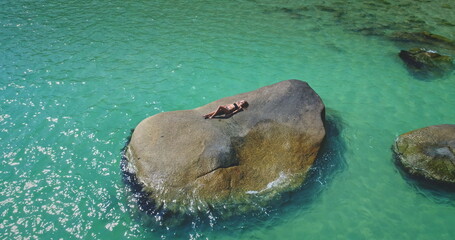 A person is lounging on a large rock in the middle of clear turquoise water. The sun shines brightly, highlighting the vibrant colors of the scene and creating a tranquil atmosphere.