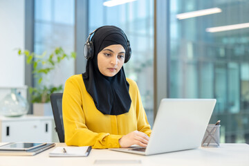 A woman wearing a hijab and headphones is working on her laptop in a modern office setting, focused intently.