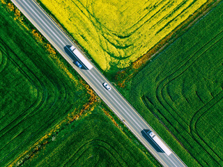 Aerial view of white trucks and cars driving on asphalt road along the green and yellow fields in Finland. Cargo delivery logistic concept