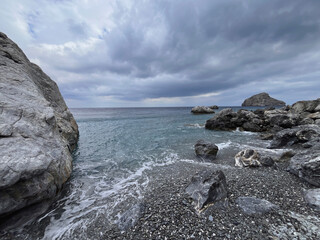 Dramatic rocky beach with black pebbles and turquoise sea on Amorgos Island, Greece – stormy sky...