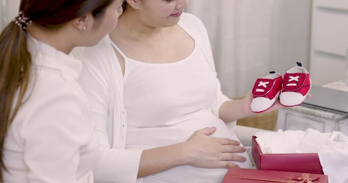 Asian pregnant woman holding tiny red baby shoes from gift box while sitting on sofa with close friend, both sharing joyful smile and excitement, enjoying beautiful moment of maternity preparation