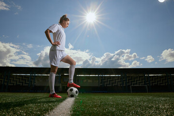 Woman footballer poses with soccer ball under sunny sky on outdoor stadium field.